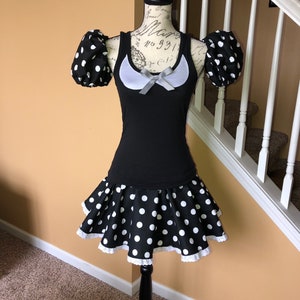 May include: A black and white polka dot dress with a white lace trim. The dress has a scoop neck and short sleeves with puffed shoulders. The dress is on a mannequin.