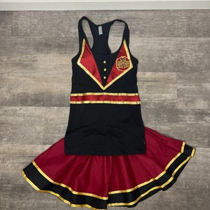 K&ouml;nnte beinhalten: Ein schwarzes Tanktop mit rotem und goldenem Besatz und einem goldenen Logo mit der Aufschrift "The Grand Floridian". Das Tanktop wird mit einem roten Rock mit schwarzem und goldenem Besatz kombiniert.