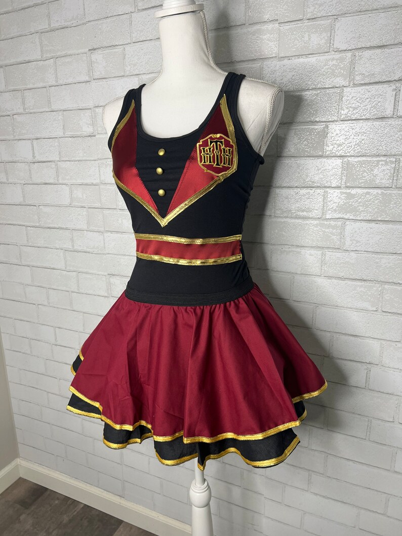 K&ouml;nnte beinhalten: Ein schwarzes und rotes &auml;rmelloses Kleid mit goldenen Verzierungen und einem goldenen gestickten Logo. Das Kleid hat einen roten Rock mit goldenen Verzierungen.