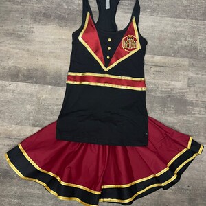 K&ouml;nnte beinhalten: Ein schwarzes Tanktop mit rotem und goldenem Besatz und einem goldenen Schlosslogo. Das Tanktop wird mit einem maroonfarbenen Rock mit schwarzem und goldenem Besatz kombiniert.