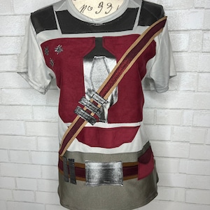 Könnte beinhalten: Ein weißes T-Shirt mit einem bedruckten Design eines Charakters, der ein rot-graues Kostüm mit einem braunen Gurt über der Brust trägt. Das Design beinhaltet ein silbernes Bruststück und einen silbernen Gürtel.