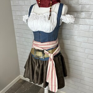 May include: A brown and white striped sash tied around a blue denim corset with a gold buckle belt. The corset has white ruffled straps and a white peasant top. A red and brown beaded necklace is worn around the neck.