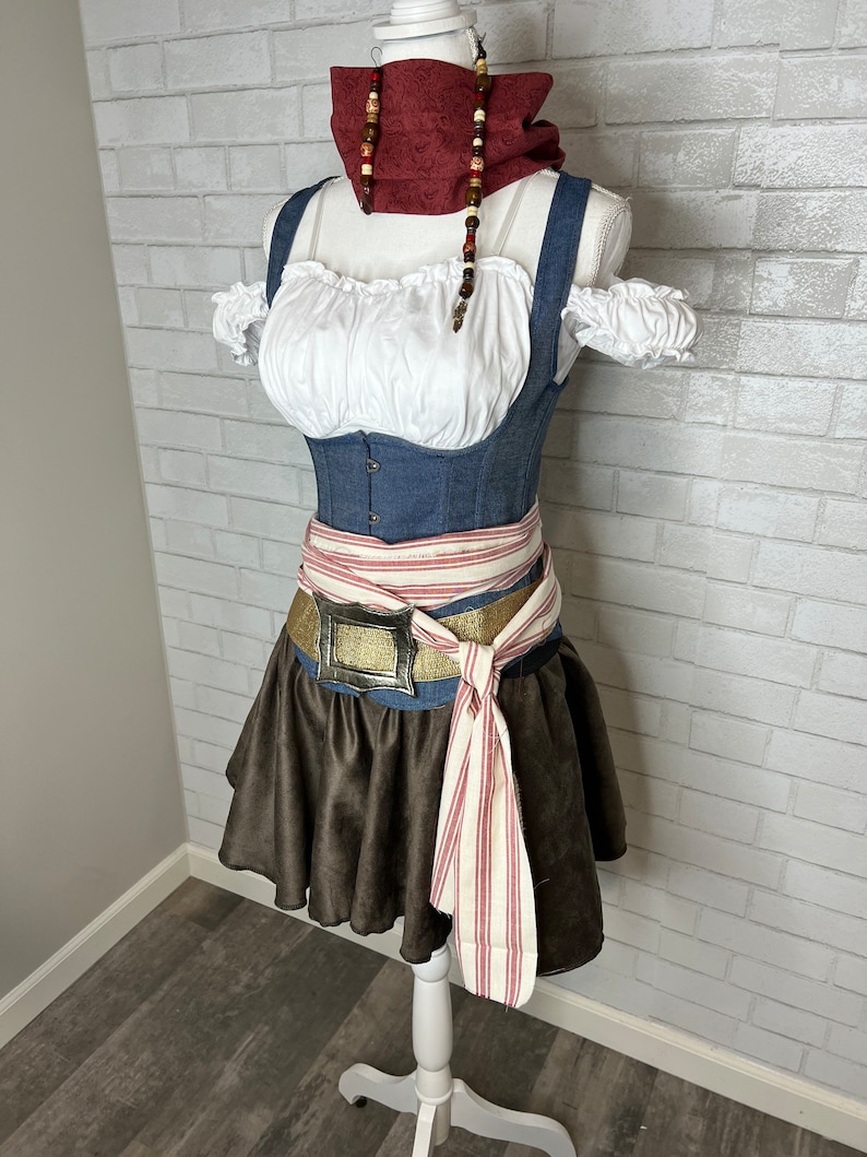 May include: A brown and white striped sash tied around a blue denim corset with a gold buckle belt. The corset has white ruffled straps and a white peasant top. A red and brown beaded necklace is worn around the neck.
