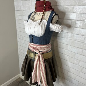 May include: A blue denim bodice with white lace trim and a brown skirt with a red and white striped sash. The bodice has a white blouse underneath and the sash is tied in a bow at the front. The mannequin is wearing a red and brown beaded necklace.