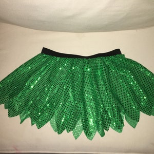 May include: A green sequined tutu skirt with a black waistband. The skirt has a jagged, leaf-like hem.