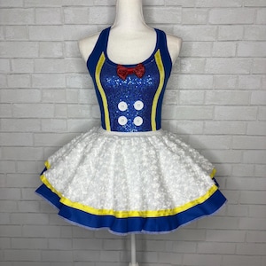 Könnte beinhalten: Ein ärmelloses Kleid mit einem blau paillettierten Oberteil, einer roten Schleife am Ausschnitt, vier weißen Knöpfen und gelben Verzierungen. Der Rock besteht aus weißem, strukturiertem Stoff mit einem gerüschten Saum.