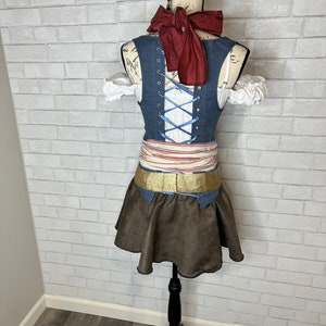 May include: A blue denim vest with a white lace-up front and a red bow tie. The vest is worn over a white blouse with lace trim. A brown skirt with a gold belt is worn over the vest.