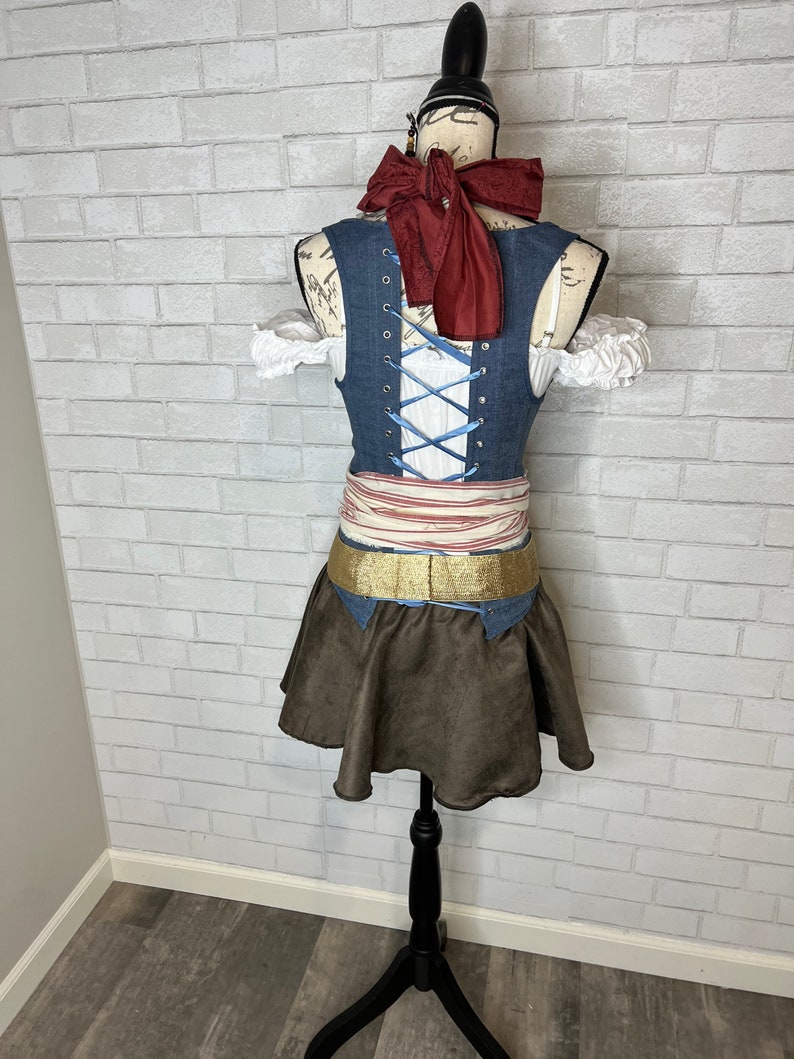 May include: A blue denim vest with a white lace-up front and a red bow tie. The vest is worn over a white blouse with lace trim. A brown skirt with a gold belt is worn over the vest.