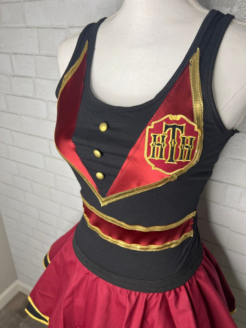 K&ouml;nnte beinhalten: Ein schwarzes und rotes Kleid mit goldenen Verzierungen. Das Kleid hat ein "Grand Floridian"-Logo auf der Vorderseite.