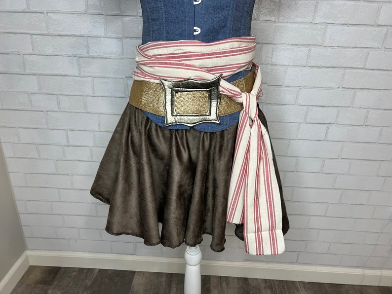 May include: A brown skirt with a gold belt and a silver buckle. The belt is tied with a white and red striped fabric.