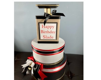 May include: A decorative birthday cake designed to resemble a perfume bottle. The cake features a black, gold, and beige color scheme, with a black bow and the text "Happy Birthday Slade". The cake is multi-tiered with red and black ribbon accents.