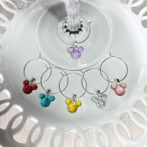 May include: Six wine glass charms featuring a sparkly Mickey Mouse design in various colors, including purple, red, teal, yellow, and silver. The charms are displayed on a white plate with a decorative edge.