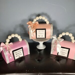 May include: Three pink, peach, and white decorative boxes shaped like purses with pearl handles and a key charm. The boxes are decorated with ribbons and rhinestones. The boxes have personalized text on the front, including "Miss Dior Christina N 30", "Tiffany's Bridal Shower", and "Gina's Fabulous 50th".