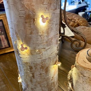 May include: A birch bark candle holder with a string of small lights and glittery Mickey Mouse-shaped ornaments. The cylindrical holder is on a wooden surface, with a second birch bark piece visible in the background.