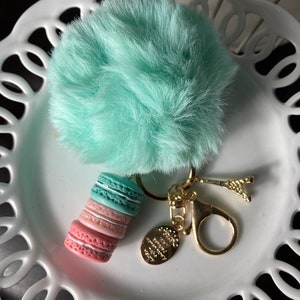 May include: A turquoise fluffy pom-pom keychain with a gold charm of a stack of macarons and a gold Eiffel Tower charm.