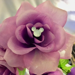 May include: Close-up of a cluster of artificial purple roses. Each rose has a silver Mickey Mouse head embellishment in the center. The petals are a soft, textured fabric. The overall theme is floral and decorative.
