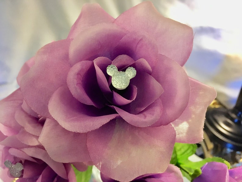 May include: Close-up of a cluster of artificial purple roses. Each rose has a silver Mickey Mouse head embellishment in the center. The petals are a soft, textured fabric. The overall theme is floral and decorative.