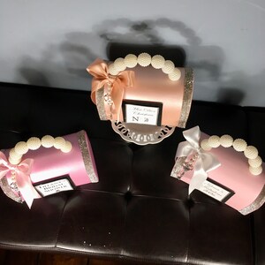 May include: Three small satin gift boxes with pearl handles and bows. The boxes are pink, peach, and white. Each box has a small tag with text that reads "Miss Dior Chrstina N 50", "Thank You Bridal Shower", and "Grandmother of the Bride".