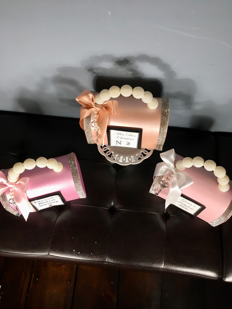 May include: Three small satin gift boxes with pearl handles and bows. The boxes are pink, peach, and white. Each box has a small tag with text that reads "Miss Dior Chrstina N 50", "Thank You Bridal Shower", and "Grandmother of the Bride".