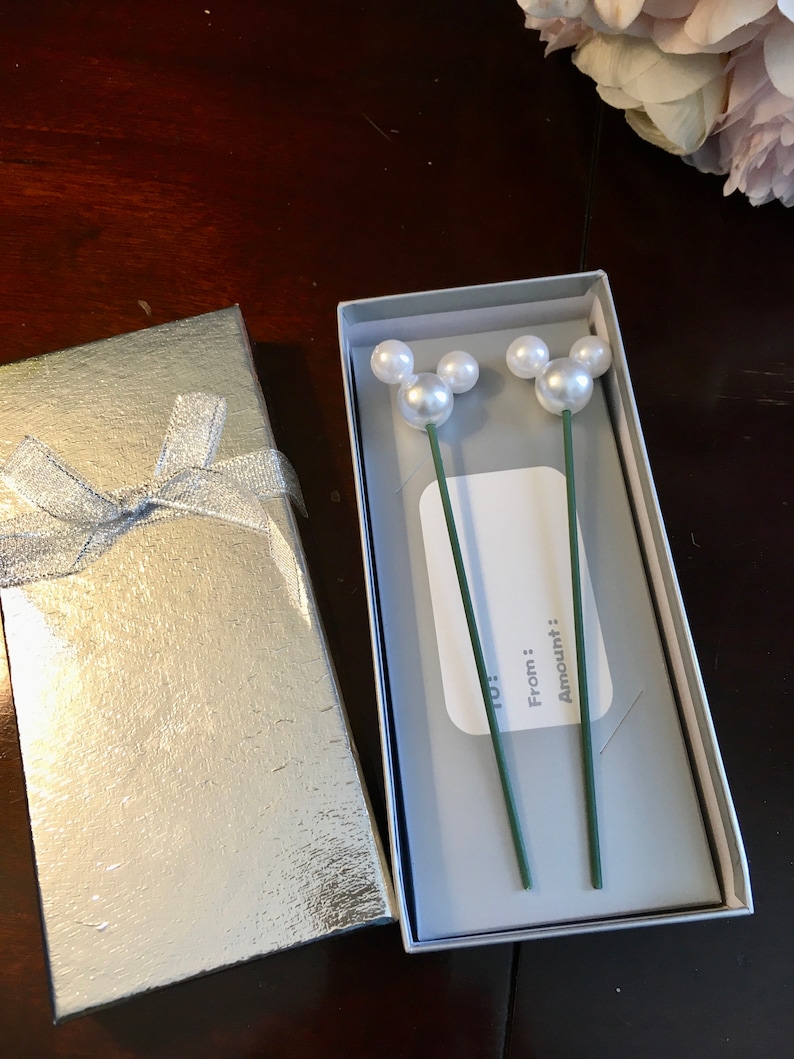 May include: A white gift box with a silver lid and a silver ribbon. The box is open and inside is a white card with the words "To:", "From:", and "Amount:" written on it. There are two green stems with white pearl Mickey Mouse heads on them.