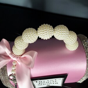 May include: Pink satin bridal shower gift box with a pearl handle and a white satin bow. The box has a black label that says "Tiffany's Bridal Shower".
