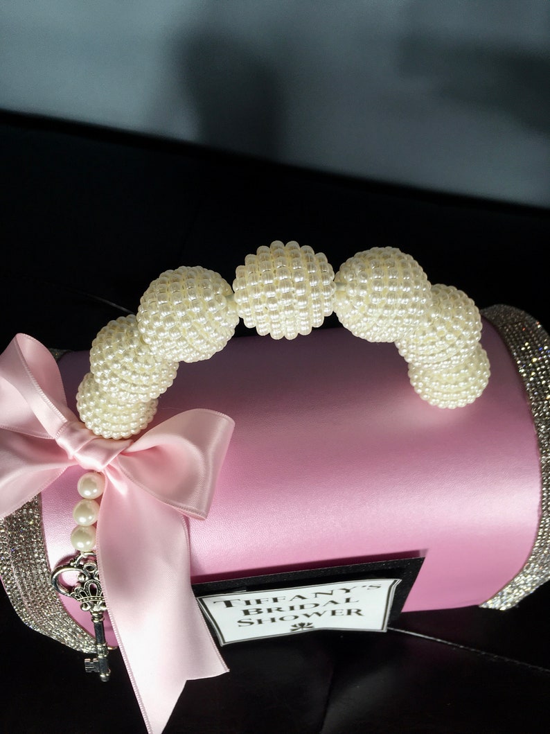 May include: Pink satin bridal shower gift box with a pearl handle and a white satin bow. The box has a black label that says "Tiffany's Bridal Shower".
