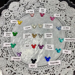 May include: A collection of glittery Mickey Mouse head-shaped embellishments in various colors, including yellow, pink, green, red, purple, and blue. Each charm is displayed on a white lace doily, with color labels.