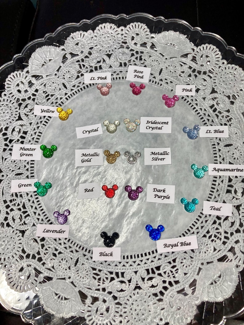 May include: A collection of glittery Mickey Mouse head-shaped embellishments in various colors, including yellow, pink, green, red, purple, and blue. Each charm is displayed on a white lace doily, with color labels.