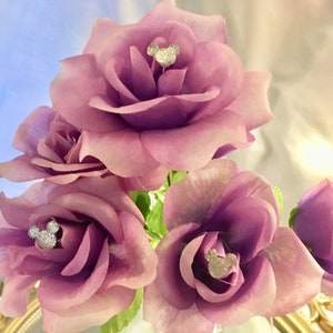 May include: A bouquet of artificial roses in shades of purple and pink. Each rose has a silver Mickey Mouse head embellishment in the center. The arrangement is set against a soft, blurred background.