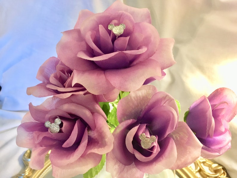 May include: A bouquet of artificial roses in shades of purple and pink. Each rose has a silver Mickey Mouse head embellishment in the center. The arrangement is set against a soft, blurred background.