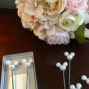 May include: A bouquet of pink and white flowers with pearl accents. The bouquet is sitting on a wooden table next to a box containing pearl Mickey Mouse head pins.