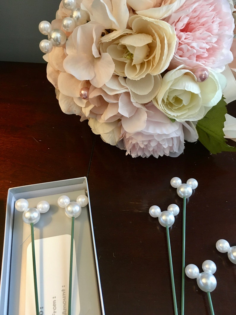 May include: A bouquet of pink and white flowers with pearl accents. The bouquet is sitting on a wooden table next to a box containing pearl Mickey Mouse head pins.