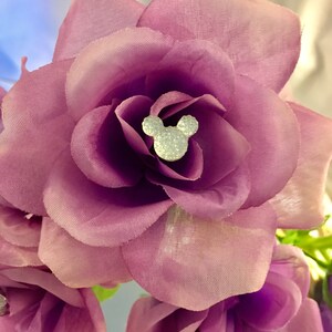 May include: Close-up of a purple fabric flower with a sparkling Mickey Mouse head in the center. The flower has multiple layers of petals and a soft texture. The background is blurred, highlighting the flower's details.