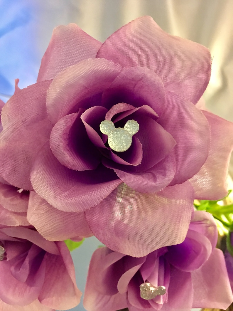 May include: Close-up of a purple fabric flower with a sparkling Mickey Mouse head in the center. The flower has multiple layers of petals and a soft texture. The background is blurred, highlighting the flower's details.