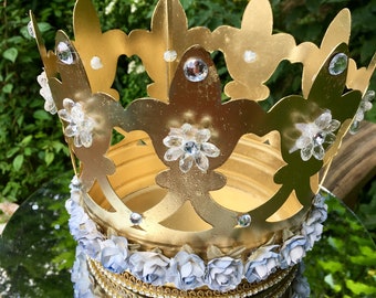 May include: A gold crown with a decorative design and blue flowers around the base. The crown is sitting on a mirrored surface.