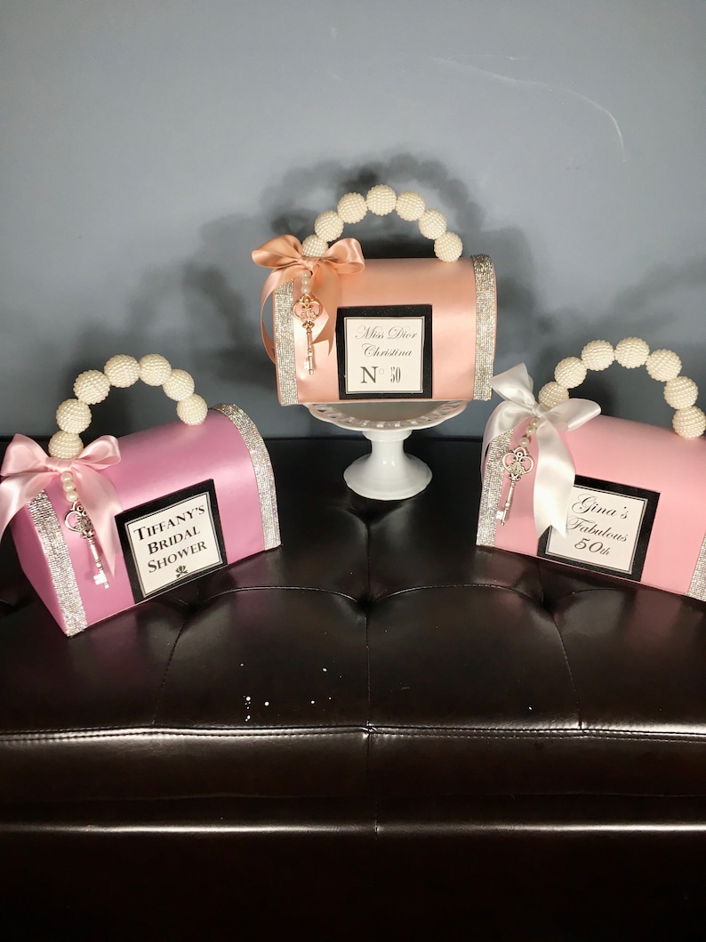 May include: Three pink, peach, and white decorative boxes shaped like purses with pearl handles and a key charm. The boxes are decorated with ribbons and rhinestones. The boxes have personalized text on the front, including "Miss Dior Christina N 30", "Tiffany's Bridal Shower", and "Gina's Fabulous 50th".