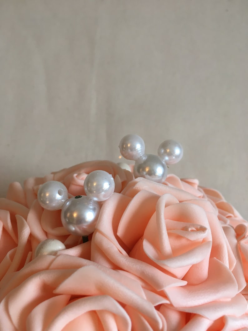 May include: A bouquet of peach-colored artificial roses with white pearl accents. The pearls are arranged in a Mickey Mouse shape.