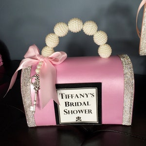 May include: Pink satin bridal shower gift box with a pearl handle, a silver key, and a white label that says "Tiffany's Bridal Shower".