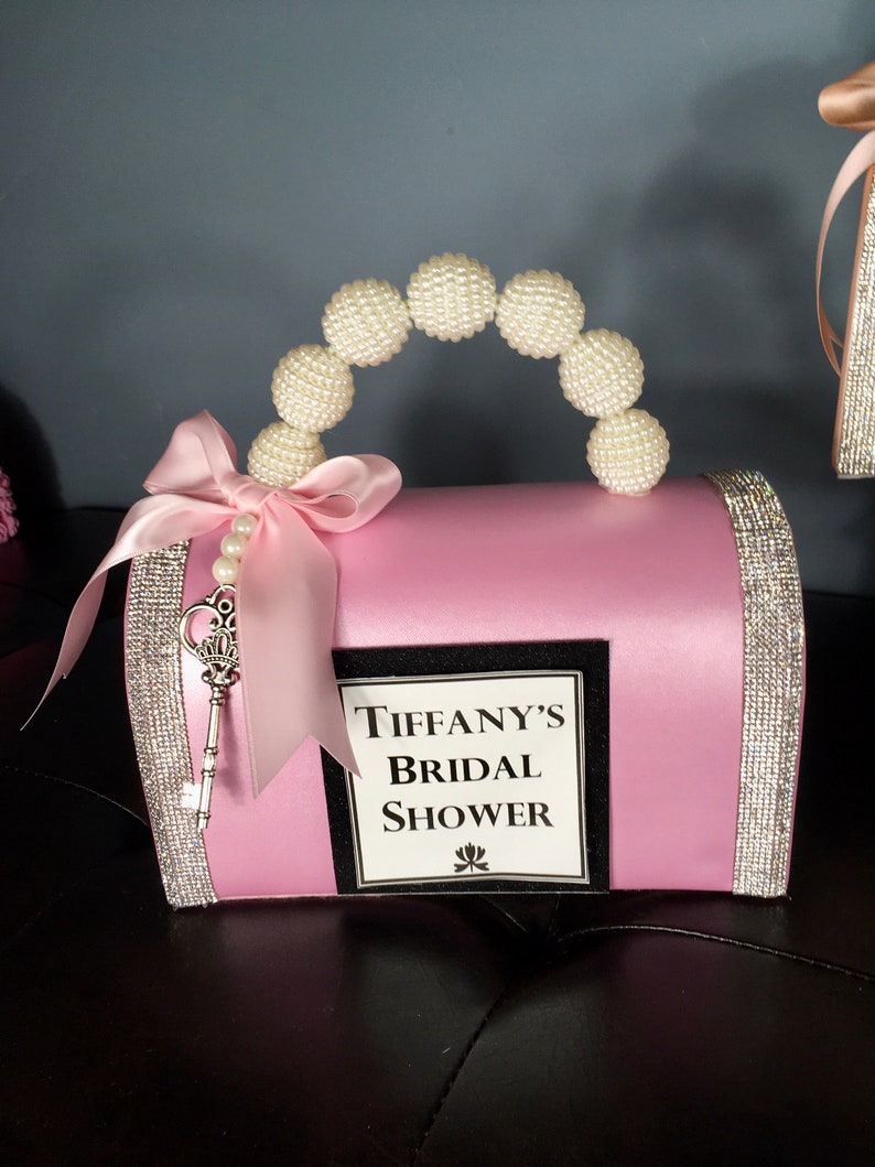 May include: Pink satin bridal shower gift box with a pearl handle, a silver key, and a white label that says "Tiffany's Bridal Shower".