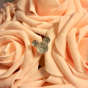 May include: A bouquet of peach-colored artificial roses with a sparkling, rhinestone-covered Mickey Mouse head embellishment. The roses have soft, velvety petals and are arranged closely together, creating a full, textured appearance. The rhinestone detail adds a touch of glamour.