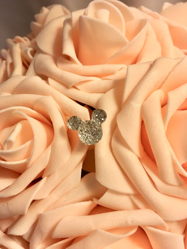 May include: A bouquet of peach-colored artificial roses with a sparkling, rhinestone-covered Mickey Mouse head embellishment. The roses have soft, velvety petals and are arranged closely together, creating a full, textured appearance. The rhinestone detail adds a touch of glamour.
