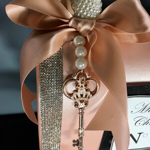 May include: A blush pink satin clutch bag with a rhinestone trim and a gold key charm. The bag has a large bow made of the same satin fabric and a pearl and rhinestone embellishment.