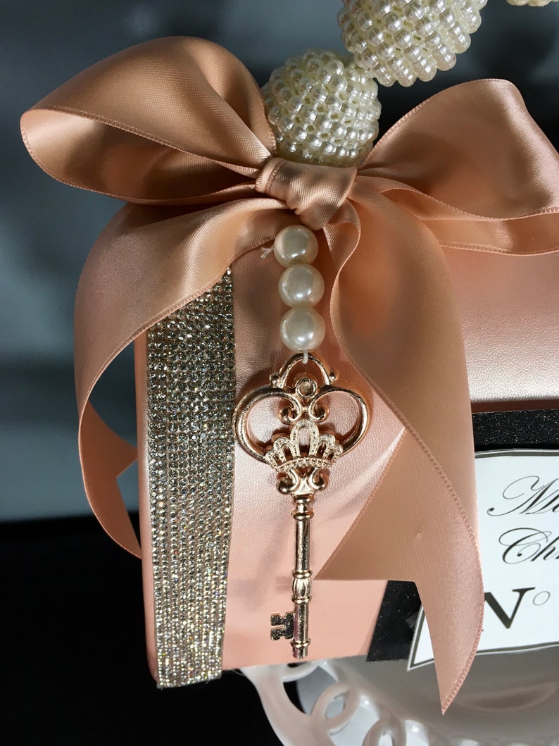 May include: A blush pink satin clutch bag with a rhinestone trim and a gold key charm. The bag has a large bow made of the same satin fabric and a pearl and rhinestone embellishment.