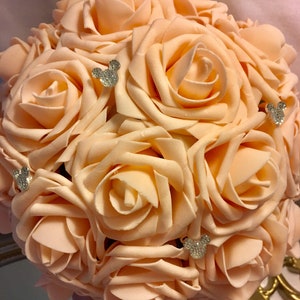 May include: A peach-colored rose bouquet with sparkling Mickey Mouse embellishments. The artificial flowers are arranged in a round shape, with a satin ribbon handle. The bouquet is a decorative item.