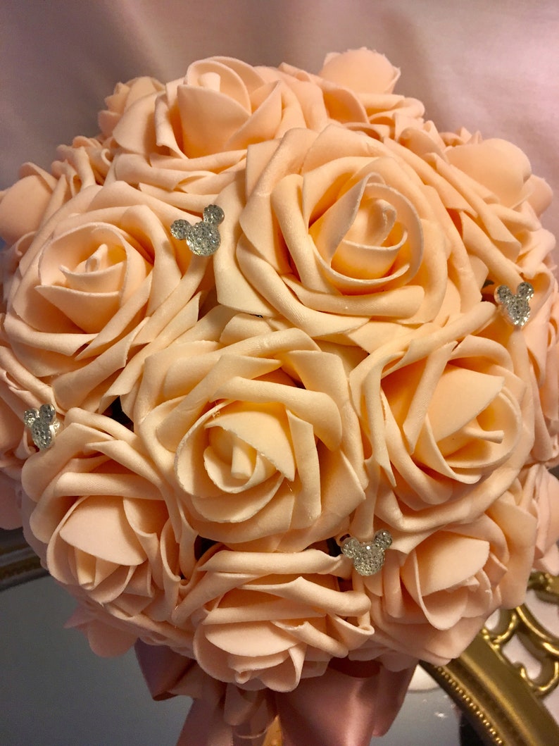 May include: A peach-colored rose bouquet with sparkling Mickey Mouse embellishments. The artificial flowers are arranged in a round shape, with a satin ribbon handle. The bouquet is a decorative item.