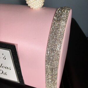 May include: A pink clutch bag with a pearl handle and a rhinestone trim. The bag has a black label with the text "It's Fabulous Ok".