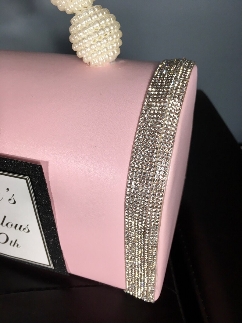 May include: A pink clutch bag with a pearl handle and a rhinestone trim. The bag has a black label with the text "It's Fabulous Ok".