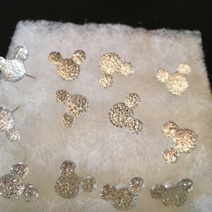 May include: A collection of silver-colored stud earrings in the shape of Mickey Mouse. The earrings are covered in small, sparkling crystals. The earrings are displayed on a white, textured surface inside a small box.