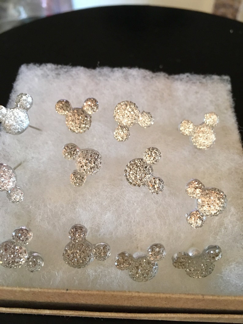 May include: A collection of silver-colored stud earrings in the shape of Mickey Mouse. The earrings are covered in small, sparkling crystals. The earrings are displayed on a white, textured surface inside a small box.
