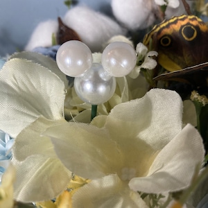 May include: A white fabric flower with a pearl Mickey Mouse head in the center. The flower is surrounded by other flowers and greenery.